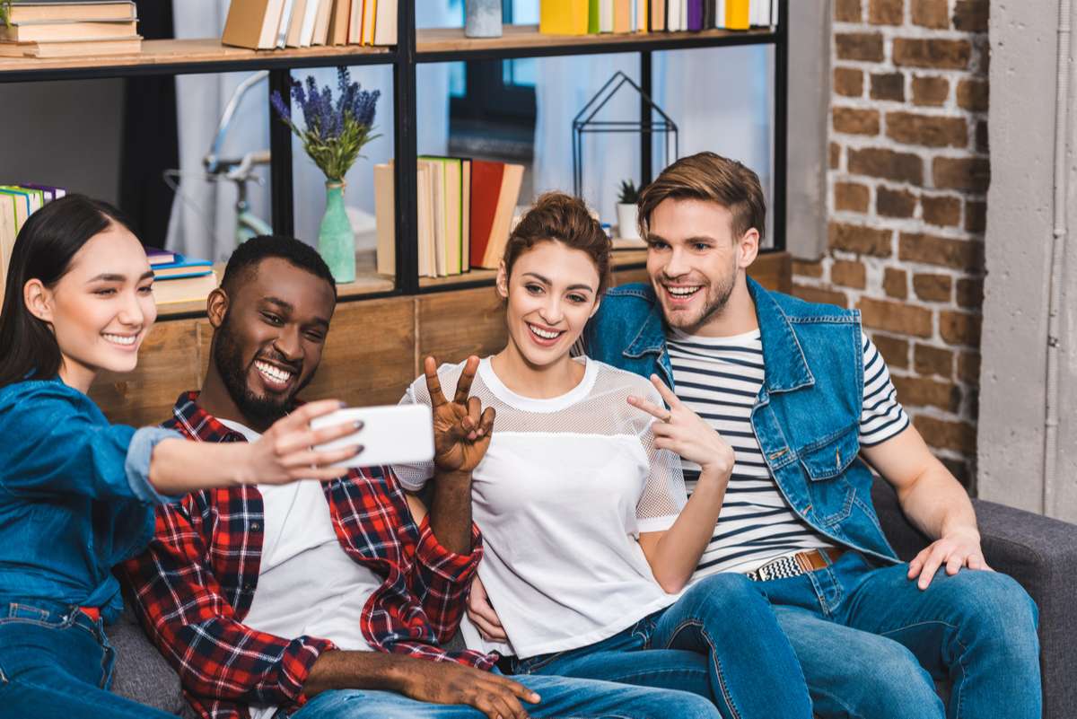 Cheerful young multiethnic friends taking selfie with smartphone while sitting on sofa (R) (S)