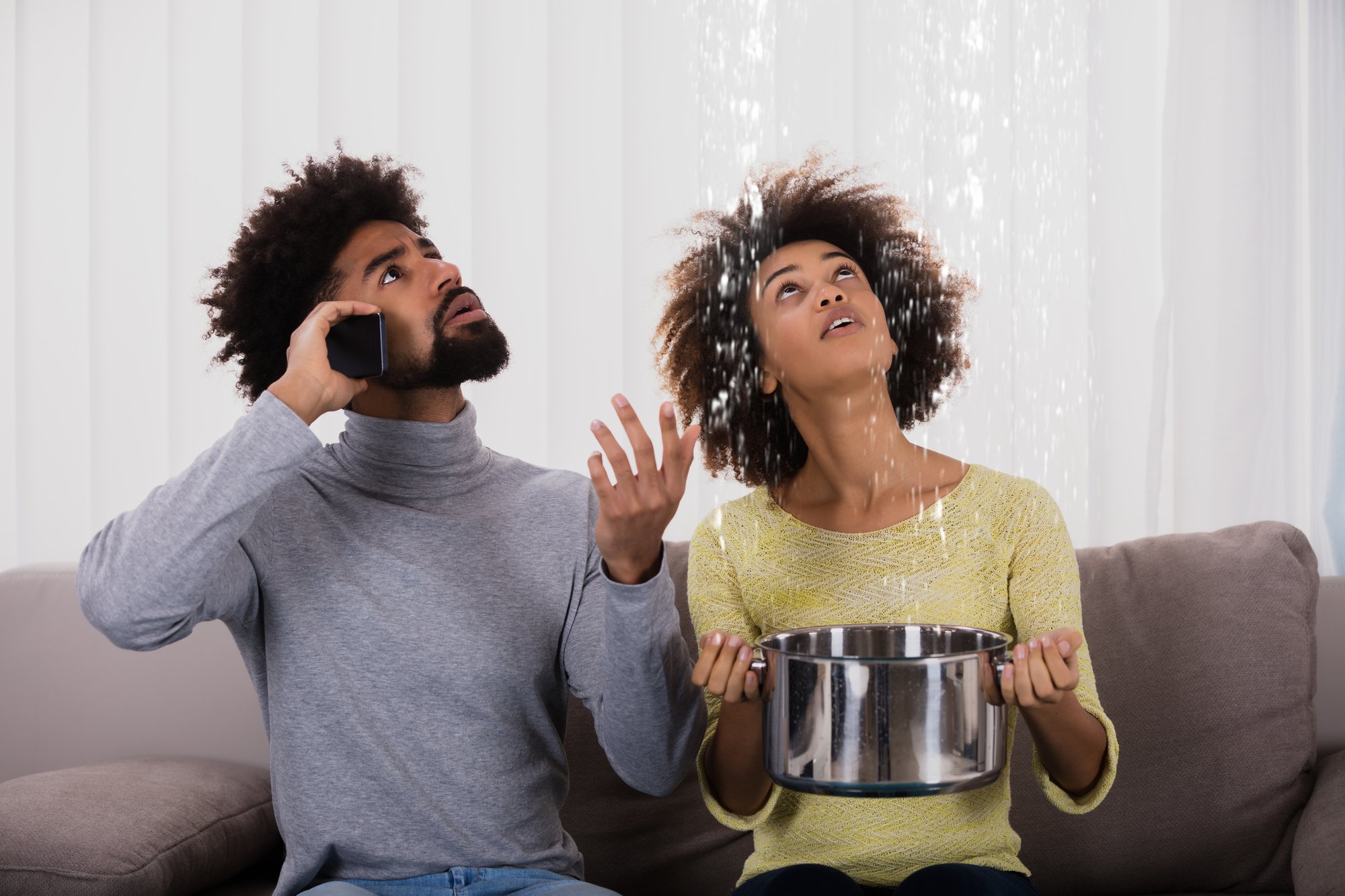 Young Man Calling Plumber While Woman Using Utensil For Collecting Water Leaking From Ceiling